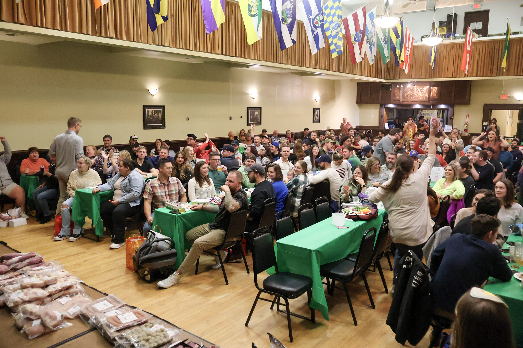 A large crowd in a huge room, mostly sitting around rectangular tables with green table clothes. Some are cheering or standing up with excitement